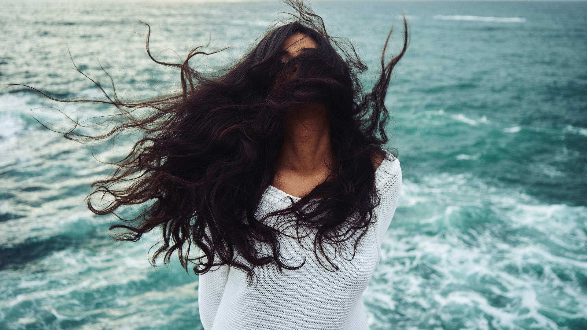Eine Frau mit dunklen langen Jahren steht im Wind am Meer, ihre Haare sind so zerzaust, dass man das Gesicht nicht erkennen kann. Eine Frau mit dunklen langen Jahren steht im Wind am Meer, ihre Haare sind so zerzaust, dass man das Gesicht nicht erkennen kann.