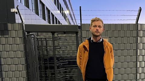 Der Abschiebebeobachter Moritz Rheinbach steht vor der Abschiebeeinrichtung der Bundespolizei am Hamburger Flughafen. Im Hintergrund ist der meterhohe Stahlzaun zu sehen. 