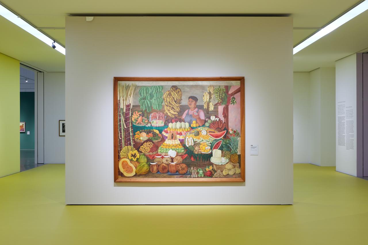 Das Gemälde "La vendedora de frutas" von Olga Costa zeigt eine Obstverkäuferin an ihrem Stand hinter vielen Sorten Obst. Sie bietet der Betrachterin eine aufgeschnittene Maracuja an.