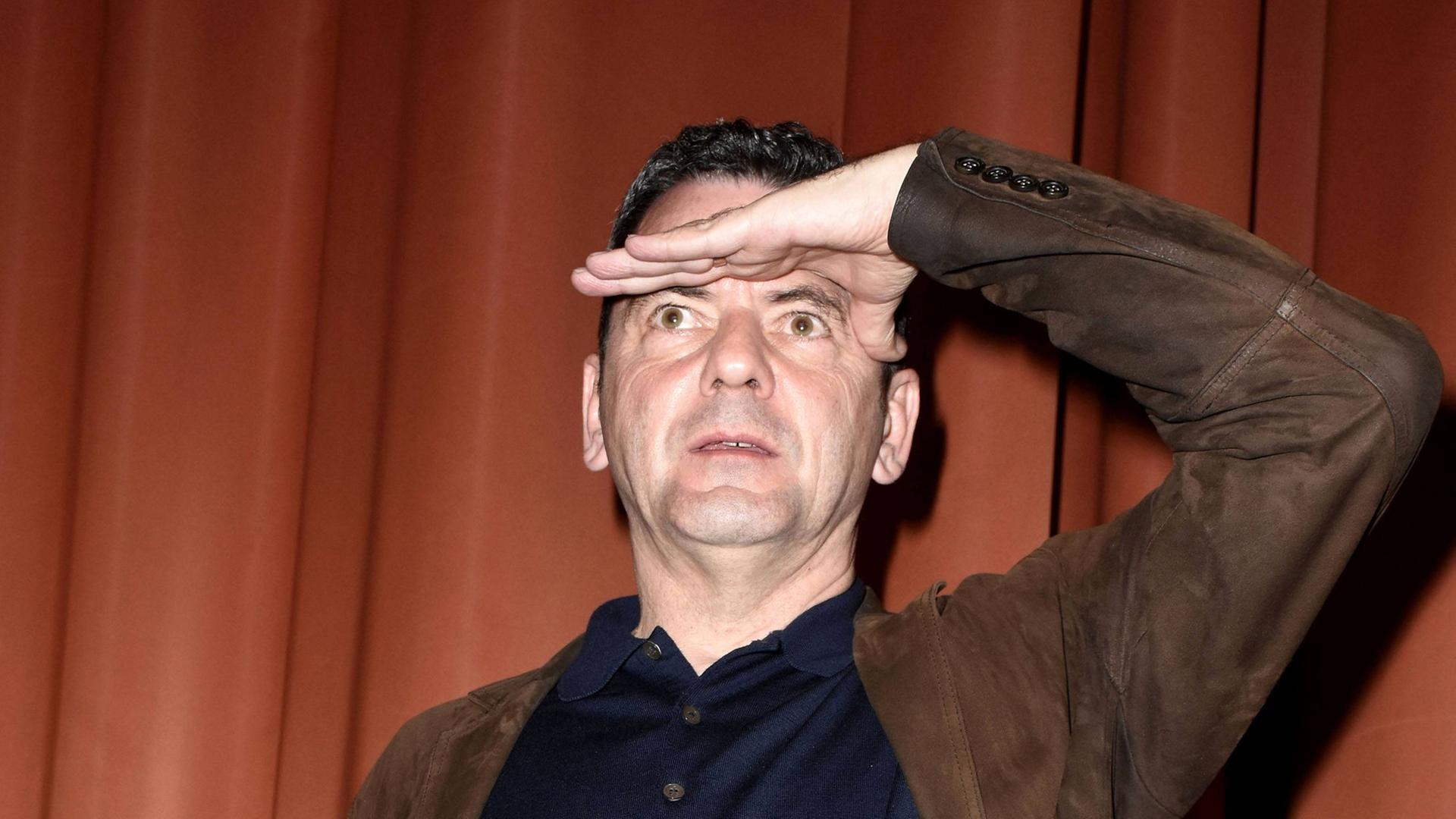 Christian Petzold steht vor einem Vorhang auf einer Bühne und schaut mit einer Handfläche an die Stirn gelegt in die Ferne. Christian Petzold steht vor einem Vorhang auf einer Bühne und schaut mit einer Handfläche an die Stirn gelegt in die Ferne.