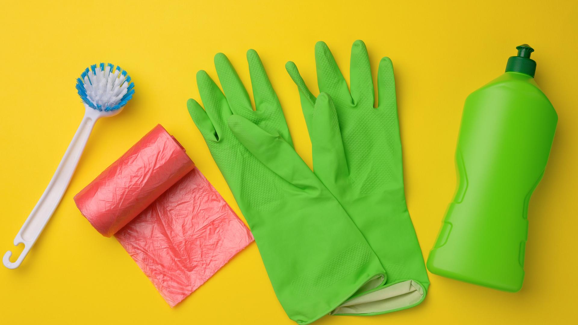 Grüne Gummihandschuhe für die Reinigung, roter Mülleimer, Plastiktütenrolle und Plastikflasche mit Reinigungsmittel auf gelbem Hintergrund (Draufsicht)