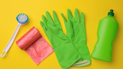 Grüne Gummihandschuhe für die Reinigung, roter Mülleimer, Plastiktütenrolle und Plastikflasche mit Reinigungsmittel auf gelbem Hintergrund (Draufsicht)