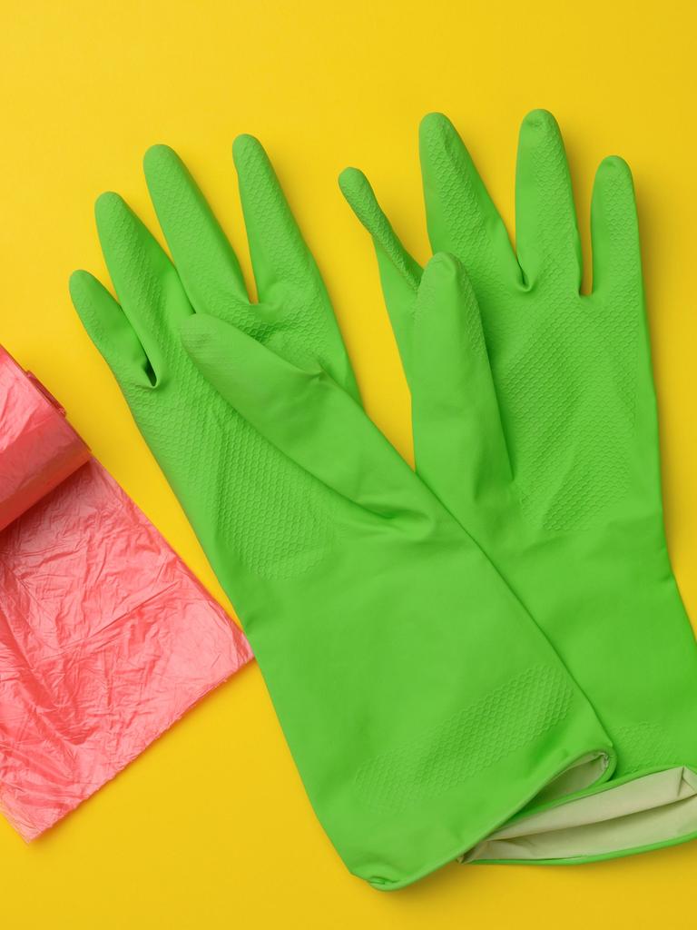 Grüne Gummihandschuhe für die Reinigung, roter Mülleimer, Plastiktütenrolle und Plastikflasche mit Reinigungsmittel auf gelbem Hintergrund (Draufsicht)