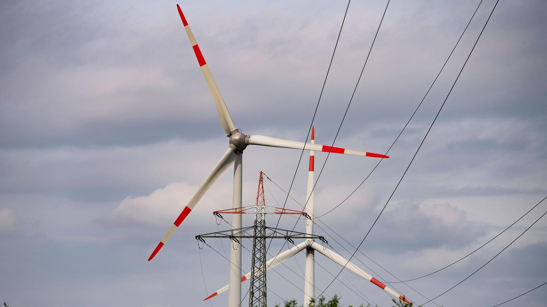 Hinter einer Hochspannungsleitung sind zwei Windräder zu sehen. Hinter einer Hochspannungsleitung sind zwei Windräder zu sehen.