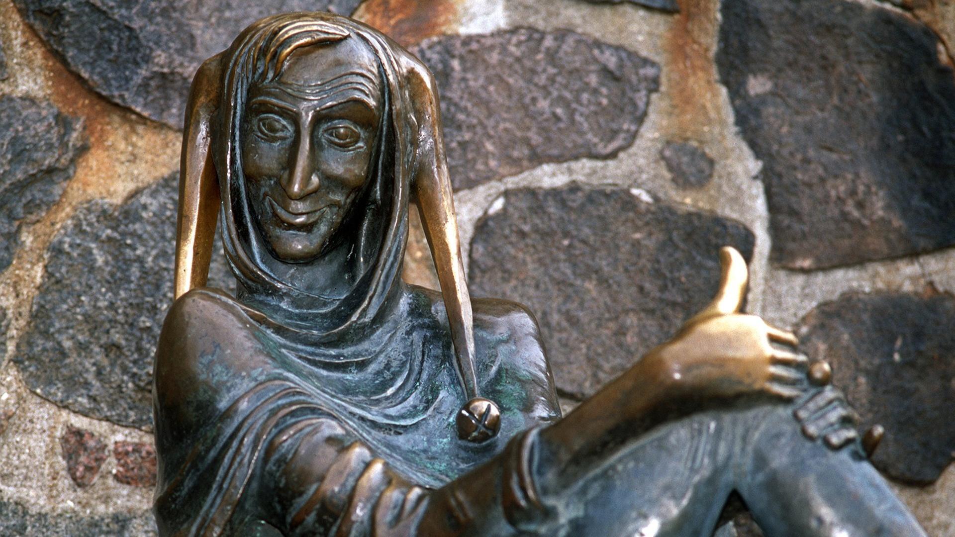 Eine Bronzeskulptur zeigt einen schelmisch grinsenden Mann mit Schellenmütze, es ist Till Eulenspiegel Eine Bronzeskulptur zeigt einen schelmisch grinsenden Mann mit Schellenmütze, es ist Till Eulenspiegel