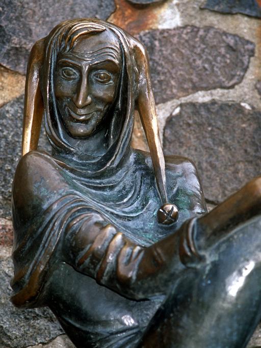 Eine Bronzeskulptur zeigt einen schelmisch grinsenden Mann mit Schellenmütze, es ist Till Eulenspiegel