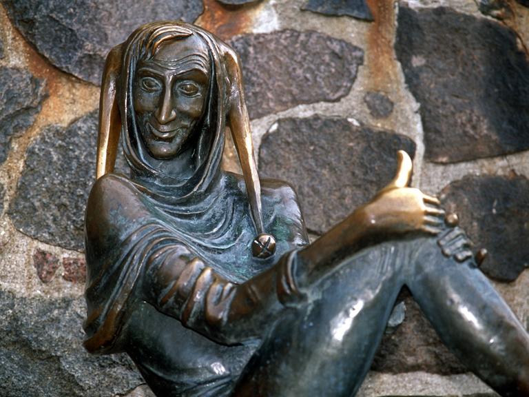 Eine Bronzeskulptur zeigt einen schelmisch grinsenden Mann mit Schellenmütze, es ist Till Eulenspiegel