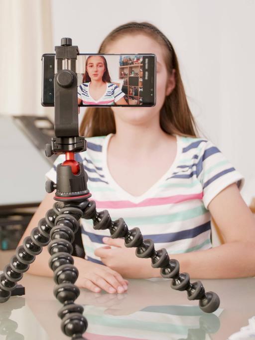 Ein Mädchen sitzt vor einer Handy-Kamera in einem Zimmer und filmt sich selbst.