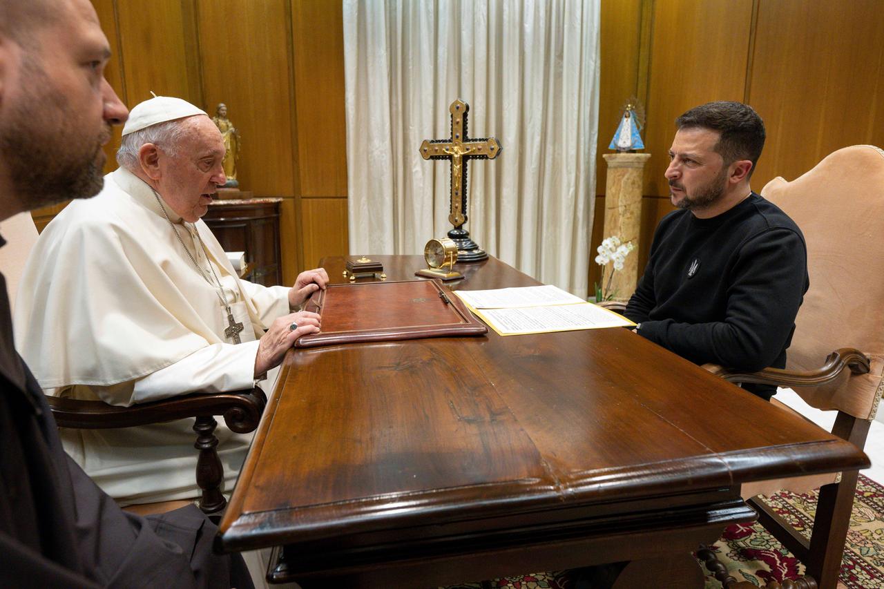Papst Franziskus sitzt gegenüber dem ukrainischen Präsidenten Wolodymyr Selenskyj an einem dunklen Holztisch. Darauf befindet sich unter anderem ein Kreuz.