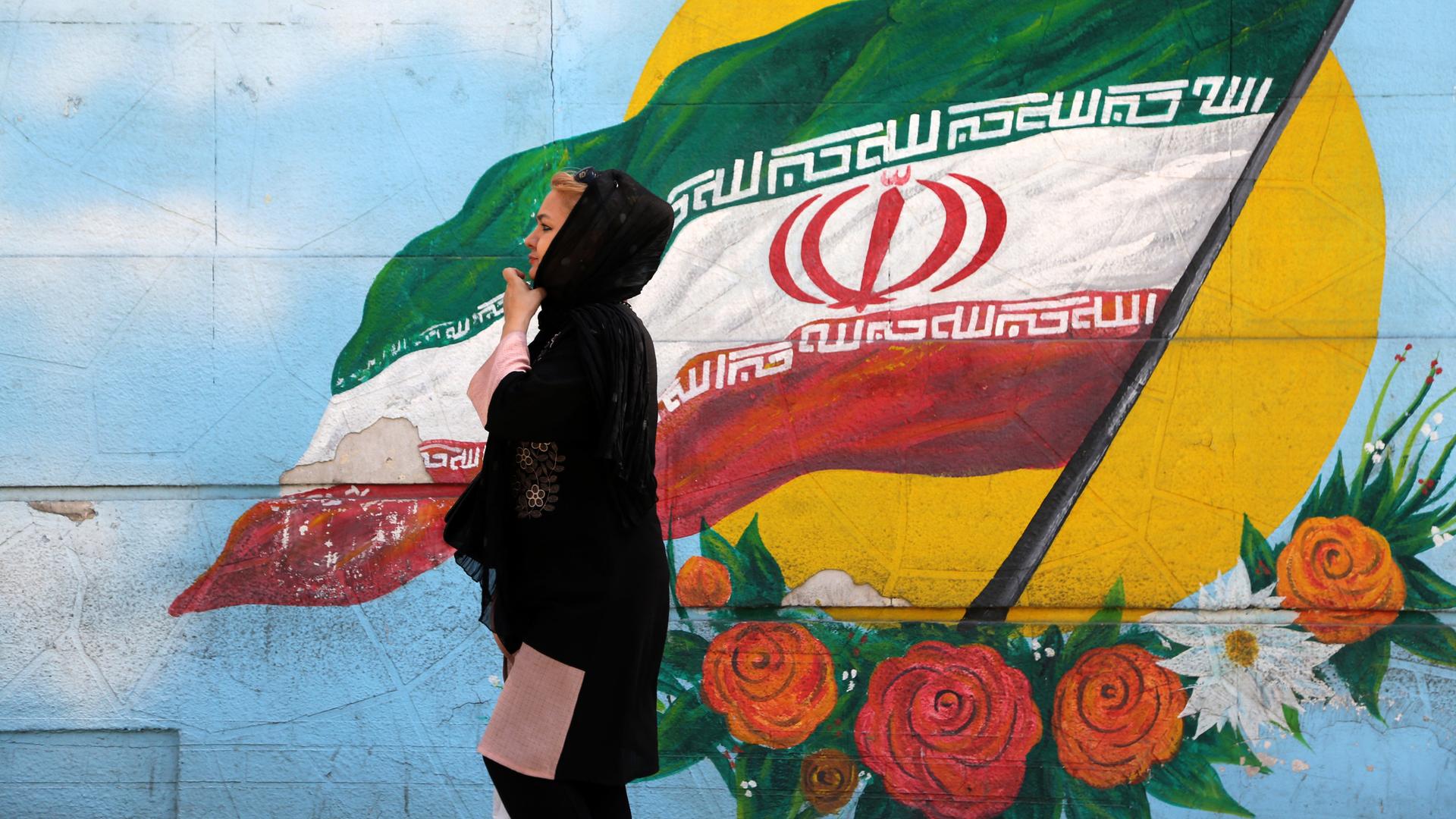 Eine Frau geht in Teheran auf der Straße, im Hintergrund ist die iranische Flagge an eine Wand gemalt.