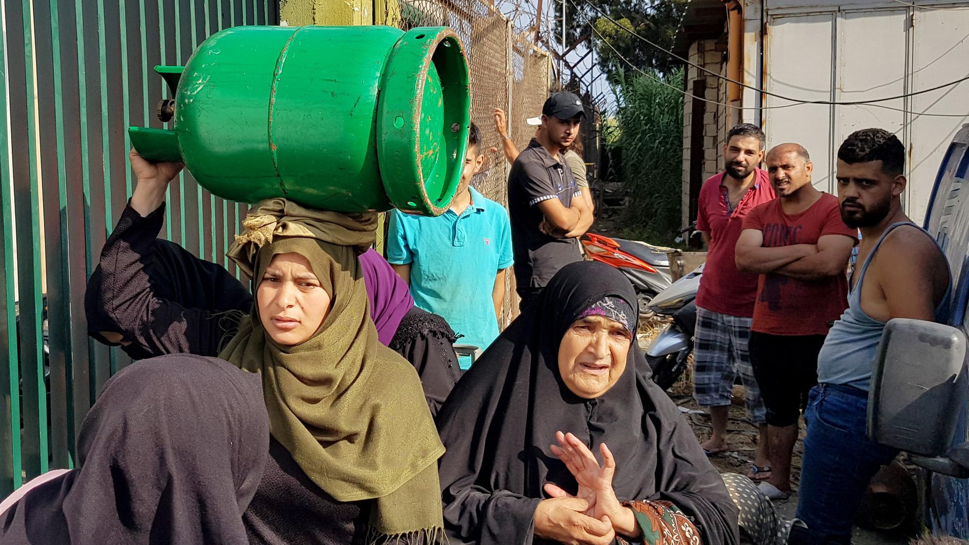 Eine Frau mit Kopftuch trägt eine grüne Gasflasche auf ihren Kopf, inmitten einer Gruppe von wartenden Frauen und Männern, die ihr nachschauen. Eine Frau mit Kopftuch trägt eine grüne Gasflasche auf ihren Kopf, inmitten einer Gruppe von wartenden Frauen und Männern, die ihr nachschauen.