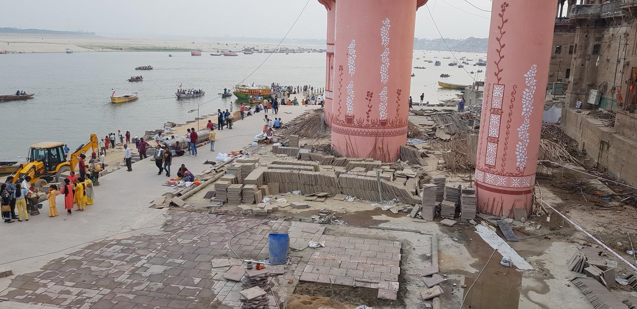 Eine Übersichtsaufnahme von den Bauarbeiten am Lalita Ghat. Auf der linken Seite ist der Ganges zu sehen, auf der rechten Seite erheben sich auf der Baustelle drei große, rosafarbene Säulen empor. Eine Übersichtsaufnahme von den Bauarbeiten am Lalita Ghat. Auf der linken Seite ist der Ganges zu sehen, auf der rechten Seite erheben sich auf der Baustelle drei große, rosafarbene Säulen empor.