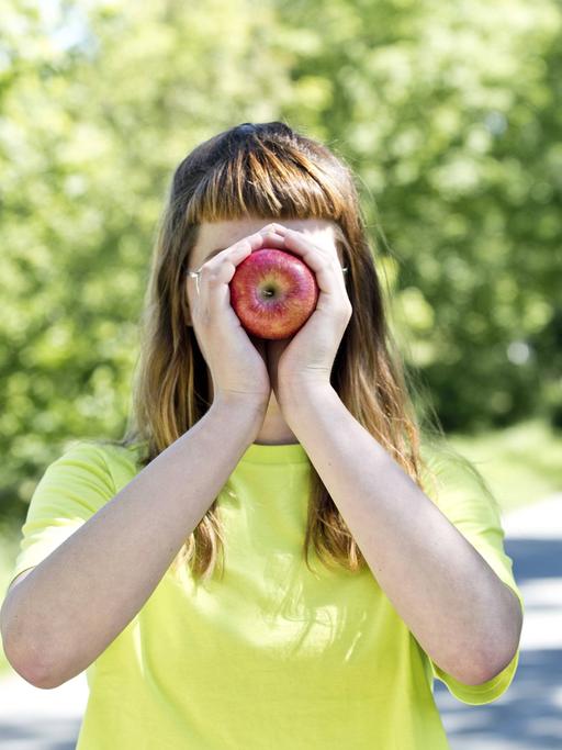 Eine junge Frau hält sich einen Apfel vor ihr Gesicht. (Symbolfoto)