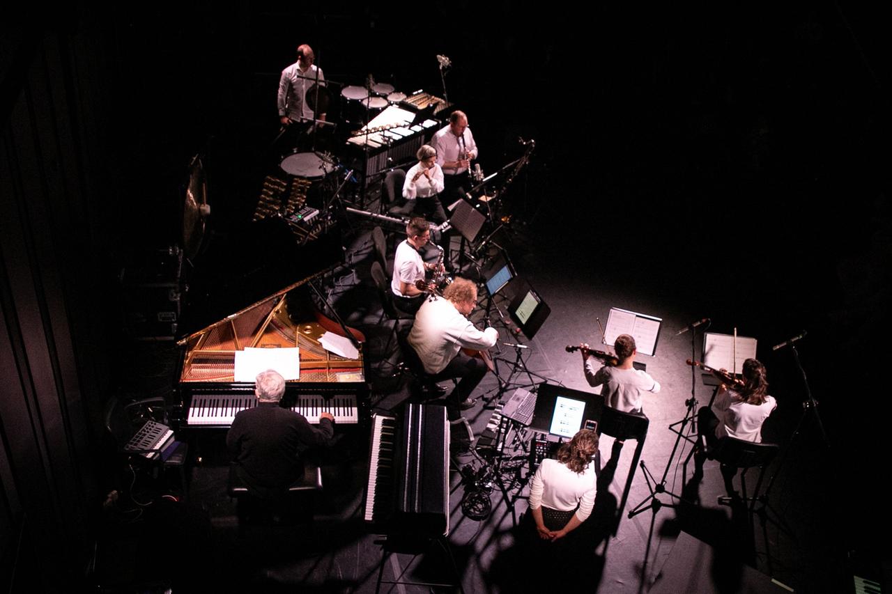 Auf dem Bild ist aus der Vogelperspektive ein Musik-Ensemble zu sehen, das umrahmt von einem schwarzen Umfeld auf einer Bühne aufspielt.