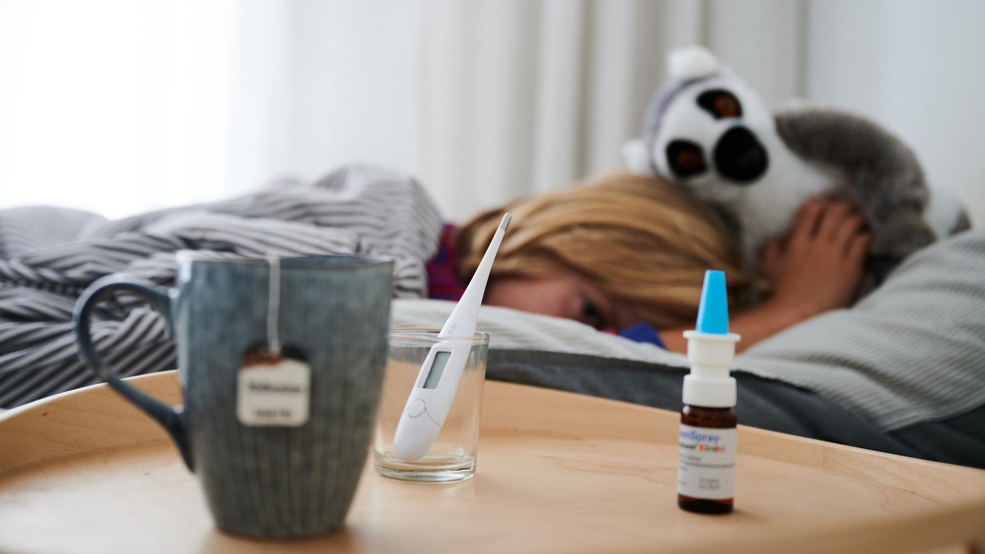 Fieberthermometer, Nasenspray und eine Tasse Tee stehen vor einem Bett, in dem ein Kind mit Kuscheltier liegt. Fieberthermometer, Nasenspray und eine Tasse Tee stehen vor einem Bett, in dem ein Kind mit Kuscheltier liegt.