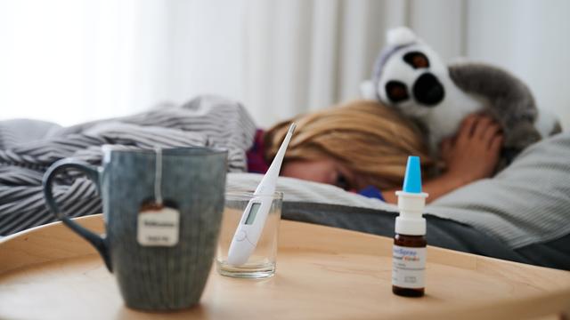 Fieberthermometer, Nasenspray und eine Tasse Tee stehen vor einem Bett, in dem ein Kind mit Kuscheltier liegt.