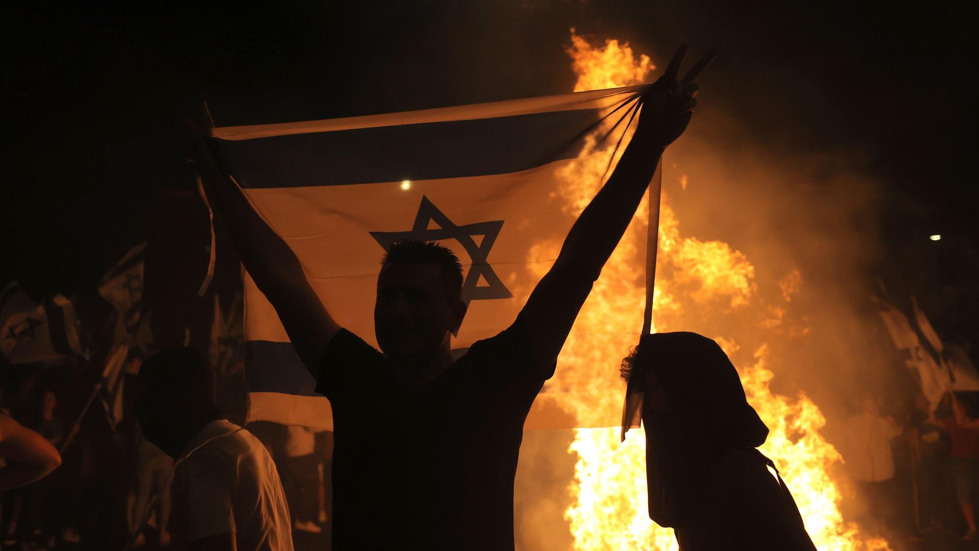 Ein Mann hält eine Israel-Flagge mit beiden Händen hoch. Hinter ihm ist ein Feuer zu sehen. 