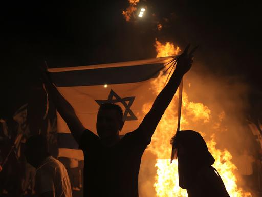 Ein Mann hält eine Israel-Flagge mit beiden Händen hoch. Hinter ihm ist ein Feuer zu sehen. 