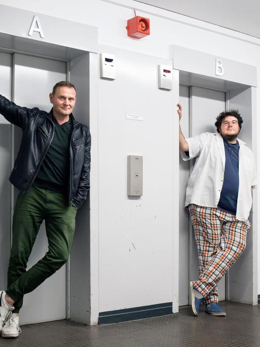 Devid Striesow und Axel Rahnisch stehen jeweils vor einer Fahrstuhltür.
