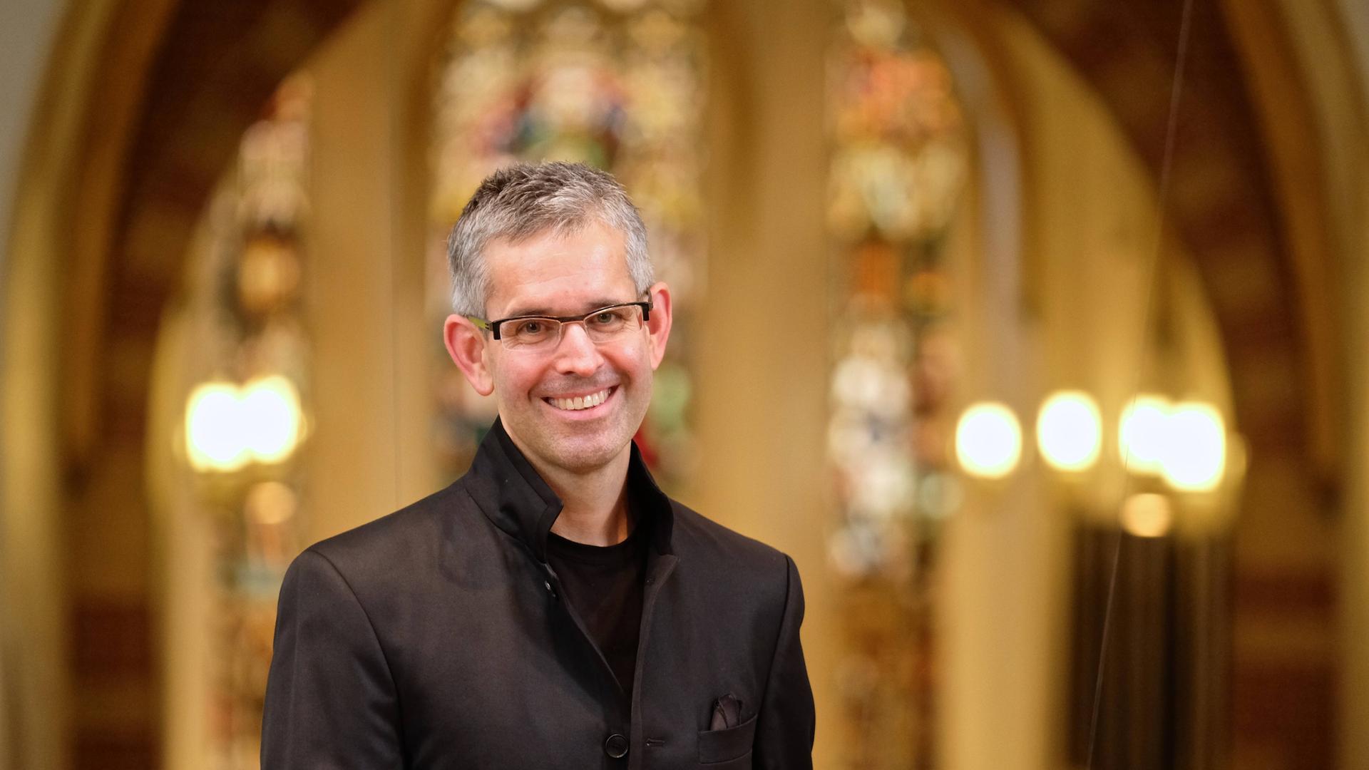 Andreas Reize steht auf einer Kirchenempore und lächelt in die Kamera. Andreas Reize steht auf einer Kirchenempore und lächelt in die Kamera.