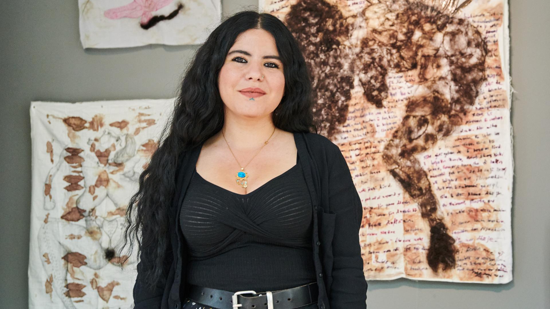 Eine schwarzhaarige junge Frau steht vor einer Wand, an der Kunstwerke hängen. Eine schwarzhaarige junge Frau steht vor einer Wand, an der Kunstwerke hängen.