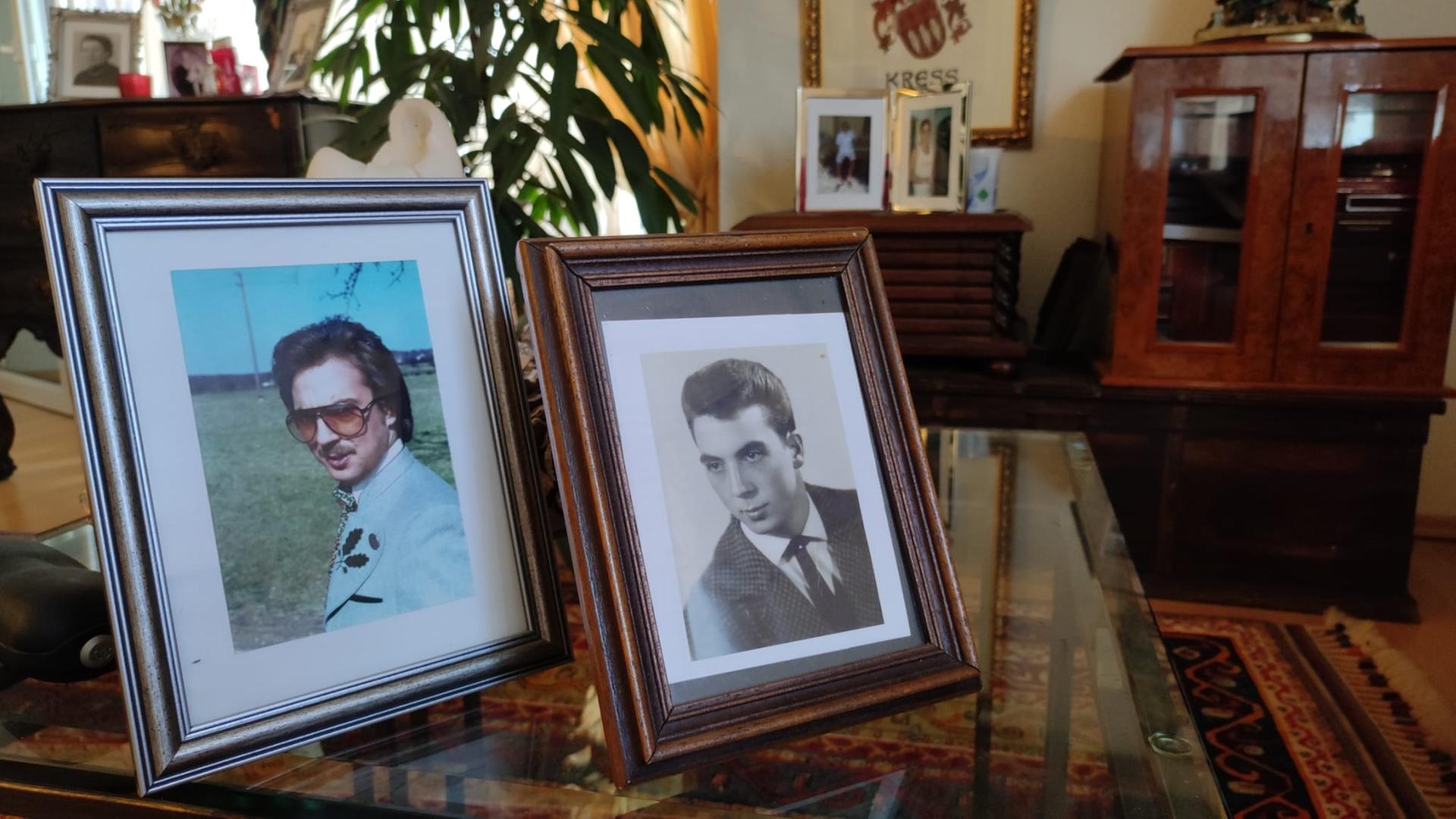 In einem Wohnzimmer stehen zwei Bilderrahmen auf einem Glastisch. Sie zeigen jeweils einen jungen Mann.