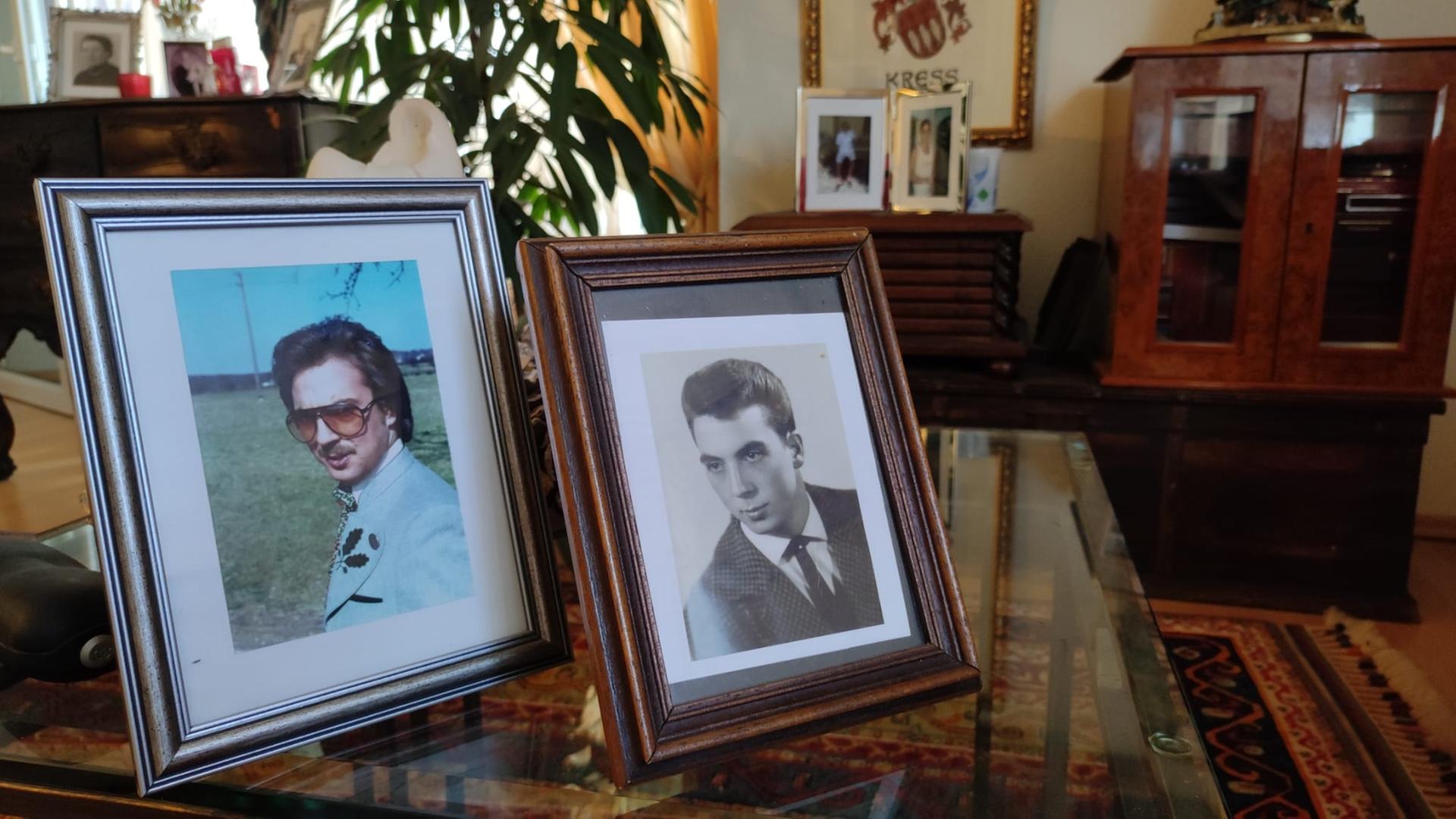 In einem Wohnzimmer stehen zwei Bilderrahmen auf einem Glastisch. Sie zeigen jeweils einen jungen Mann. In einem Wohnzimmer stehen zwei Bilderrahmen auf einem Glastisch. Sie zeigen jeweils einen jungen Mann.