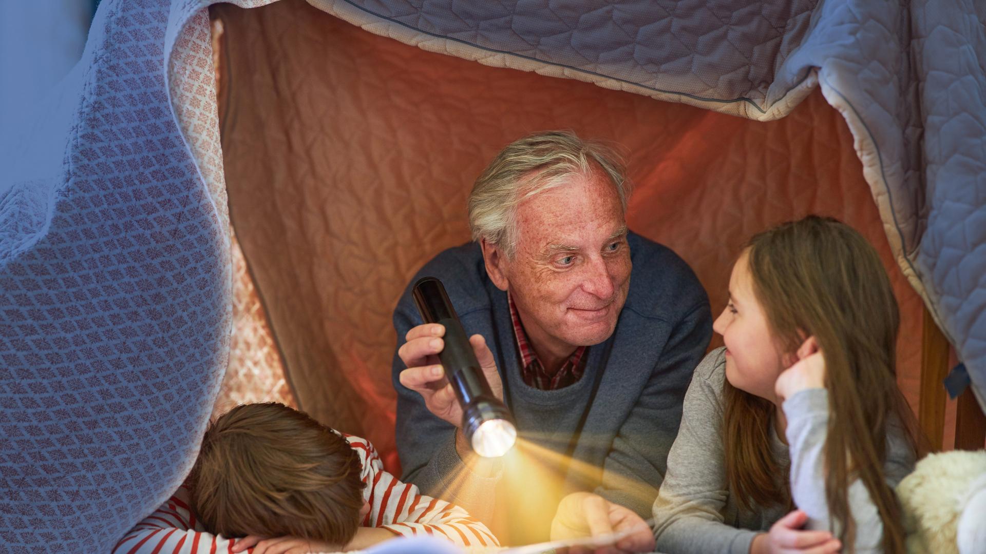 Ein Großvater liest seinen Enkeln unter einer Zeltdecke zu Hause vor 