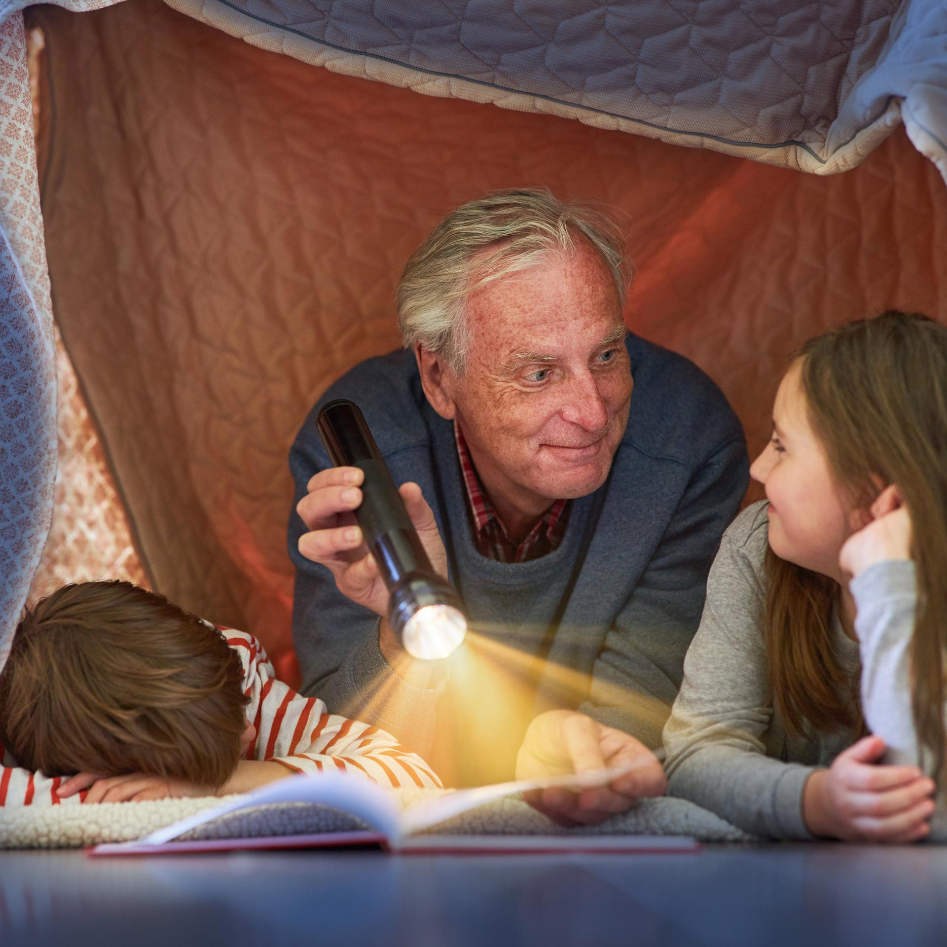 Ein Großvater liest seinen Enkeln unter einer Zeltdecke zu Hause vor 