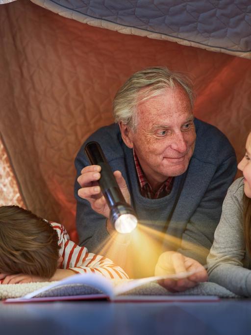 Ein Großvater liest seinen Enkeln unter einer Zeltdecke zu Hause vor 