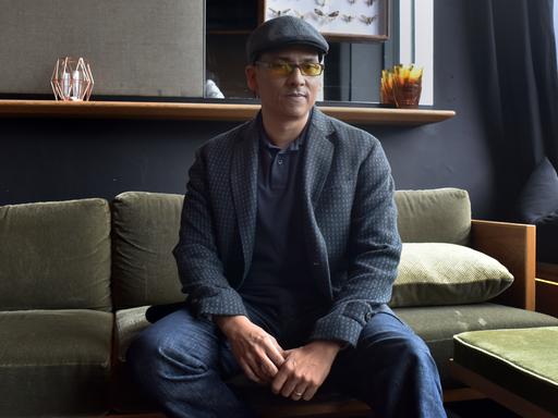 Der Sänger Xavier Naidoo sitzt auf einem Sofa und blickt in die Kamera. Er trägt eine Mütze und eine Brille mit getönten Gläsern.