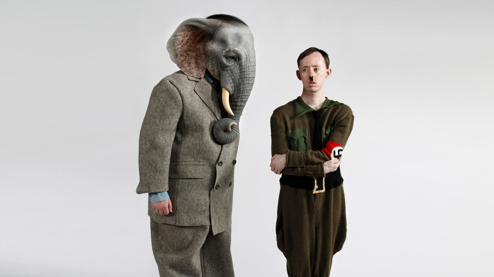 Ein Schauspieler mit einer Elefantenkopfmaske und ein zweiter, der Hitler darstellt, stehen auf einer Bühne. Ein Schauspieler mit einer Elefantenkopfmaske und ein zweiter, der Hitler darstellt, stehen auf einer Bühne.