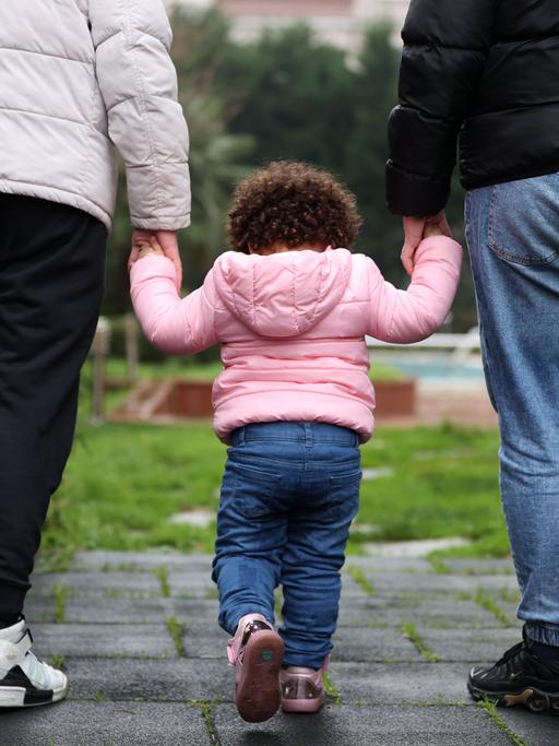 Ein kleines Kind mit lockigem Haar und rosa Anorak läuft an der Hand der Eltern über eine Wiese. 