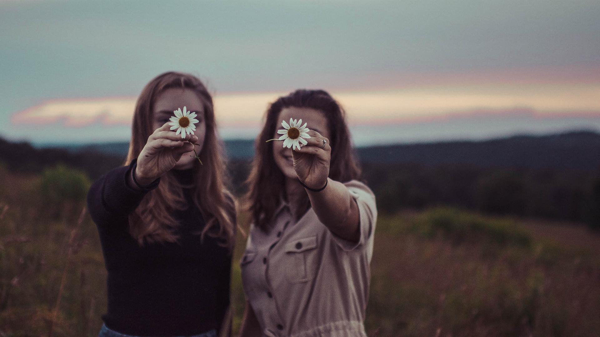 Zwei Frauen stehen vor einer wild romantischen Landschaft mit untergehender Sonne und halten lachend jeweils eine weiße Blume so vor das Gesicht, dass nur die Blumen scharf erscheinen. Zwei Frauen stehen vor einer wild romantischen Landschaft mit untergehender Sonne und halten lachend jeweils eine weiße Blume so vor das Gesicht, dass nur die Blumen scharf erscheinen.