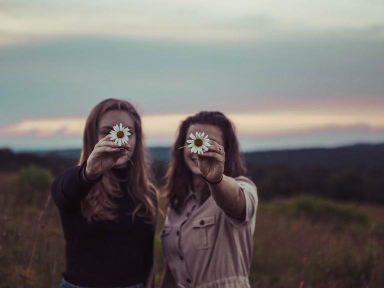 Zwei Frauen stehen vor einer wild romantischen Landschaft mit untergehender Sonne und halten lachend jeweils eine weiße Blume so vor das Gesicht, dass nur die Blumen scharf erscheinen.