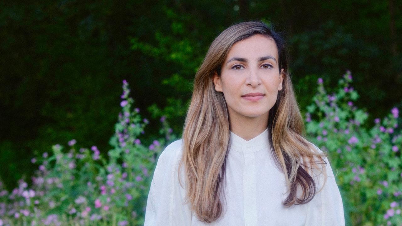 Die Aktivistin Hila Latifi steht in einem weißen Kleid auf einer Wiese und lässt beide Arme am Körper herunterhängen. Sie hat langes braunes Haar und bllickt in die Kamera. Die Aktivistin Hila Latifi steht in einem weißen Kleid auf einer Wiese und lässt beide Arme am Körper herunterhängen. Sie hat langes braunes Haar und bllickt in die Kamera.