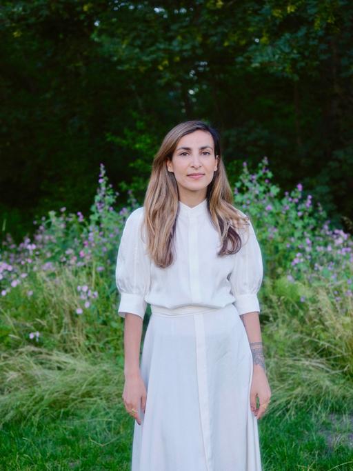 Die Aktivistin Hila Latifi steht in einem weißen Kleid auf einer Wiese und lässt beide Arme am Körper herunterhängen. Sie hat langes braunes Haar und bllickt in die Kamera.