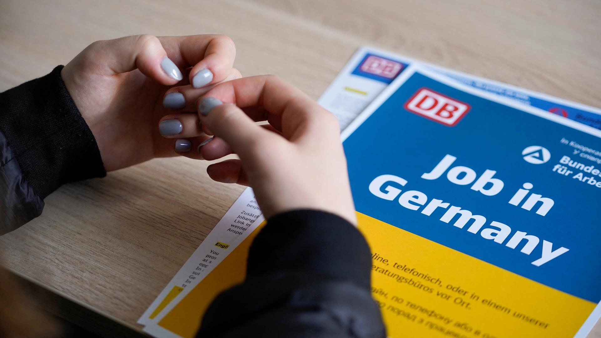 Die Hände einer Frau mit taubenblauem Nagellack liegen auf einem Informationsblatt der Deutschen Bahn, es ist in den ukrainischen Nationalfarben Blau und Gelb gehalten und trägt die Aufschrift "Job in Germany". Die Hände einer Frau mit taubenblauem Nagellack liegen auf einem Informationsblatt der Deutschen Bahn, es ist in den ukrainischen Nationalfarben Blau und Gelb gehalten und trägt die Aufschrift "Job in Germany".