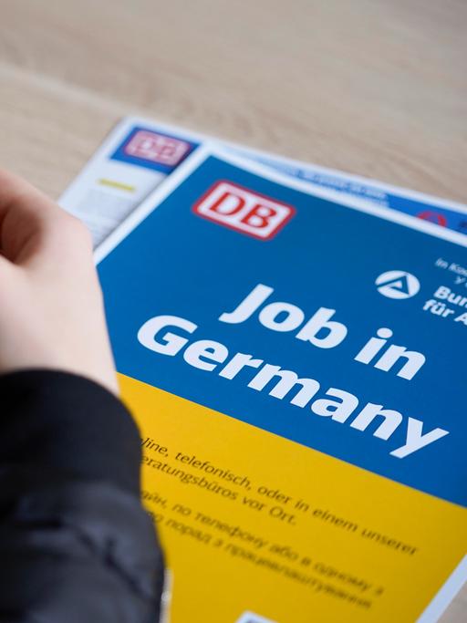 Die Hände einer Frau mit taubenblauem Nagellack liegen auf einem Informationsblatt der Deutschen Bahn, es ist in den ukrainischen Nationalfarben Blau und Gelb gehalten und trägt die Aufschrift "Job in Germany".