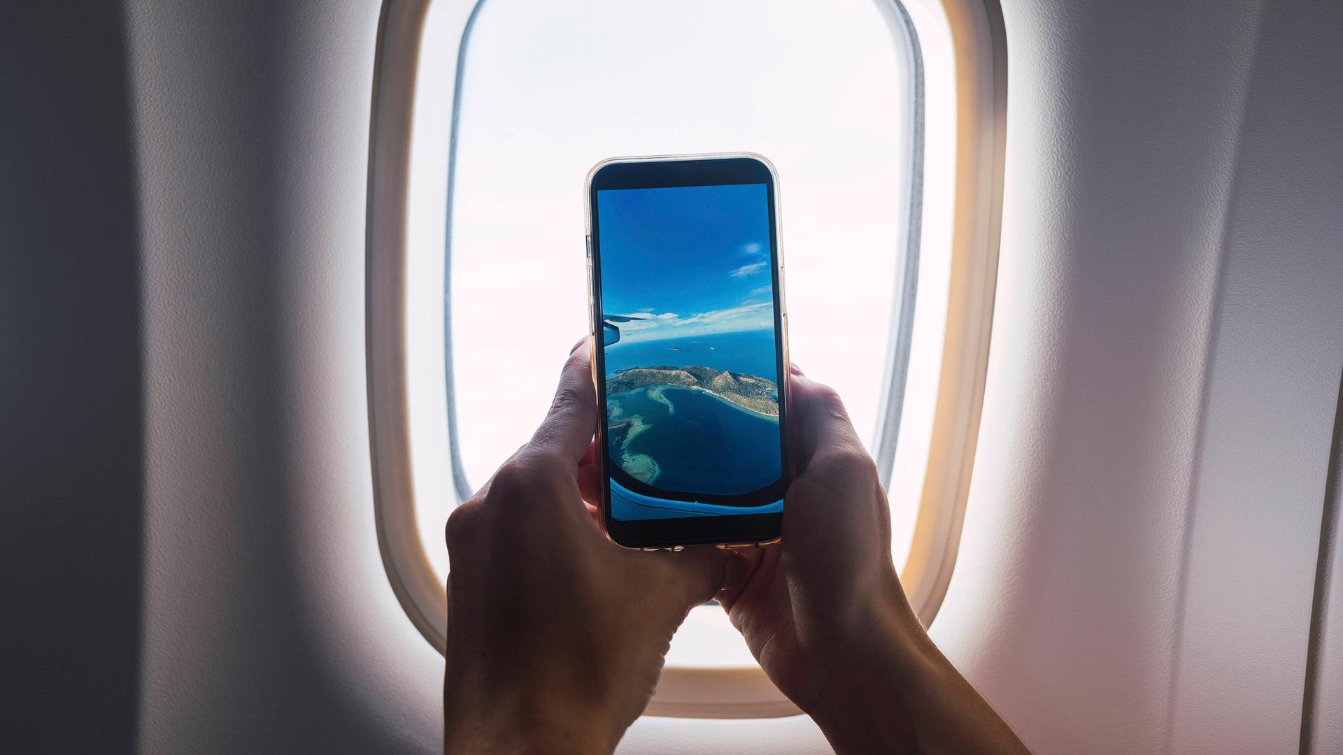 Ein Mann, von dem man nur die Hände sieht, macht mit dem Smartphone ein Foto durch das Fenster eines Flugzeugs. Ein Mann, von dem man nur die Hände sieht, macht mit dem Smartphone ein Foto durch das Fenster eines Flugzeugs.