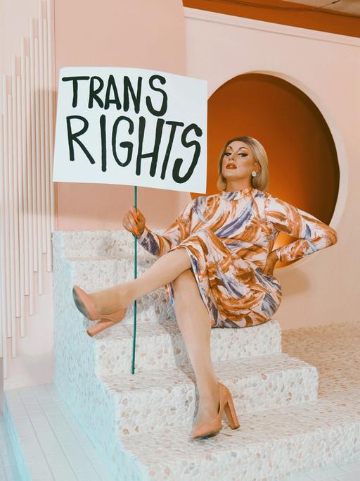 Die Dragqueen und Politikerin Maebe A. Girl mit einem Schild auf dem "Trans Rights" steht vor einer orangefarbenen Kulisse.
