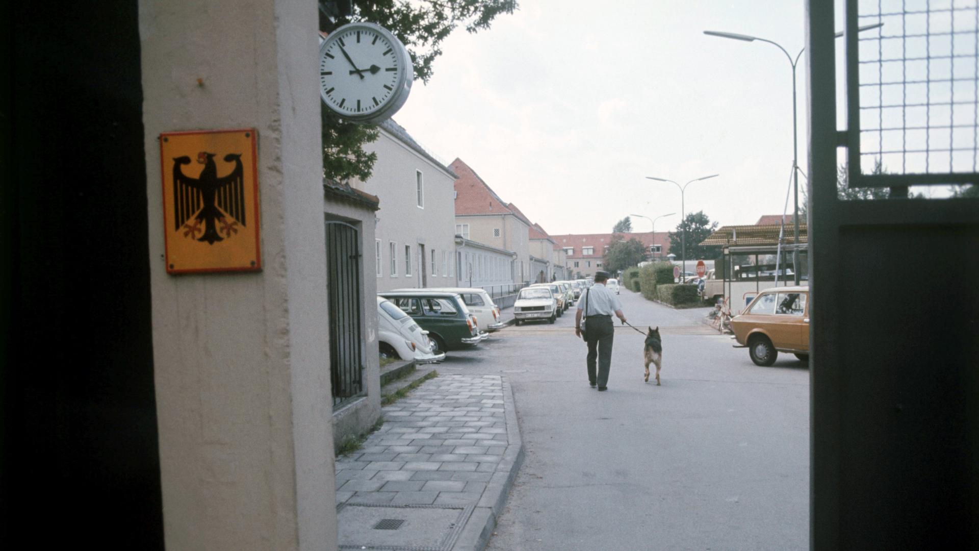Durch ein Eingangstor sieht man einen Polizeibeamten mit Hund von hinten auf einer Aufnahme des Gelände des Bundesnachrichtendienstes in Pullach um 1960/1970.