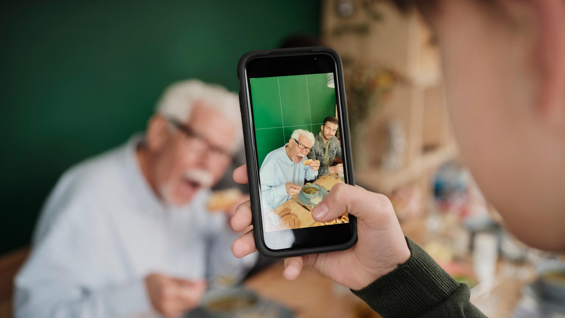 Ein Kind fotografiert zwei erwachsene Personen, die essend am Tisch sitzen. Die Szene ist unscharf, das Bild auf dem Smartphone-Display ist scharf. Ein Kind fotografiert zwei erwachsene Personen, die essend am Tisch sitzen. Die Szene ist unscharf, das Bild auf dem Smartphone-Display ist scharf.
