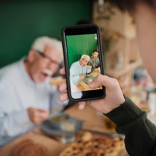 Ein Kind fotografiert zwei erwachsene Personen, die essend am Tisch sitzen. Die Szene ist unscharf, das Bild auf dem Smartphone-Display ist scharf.