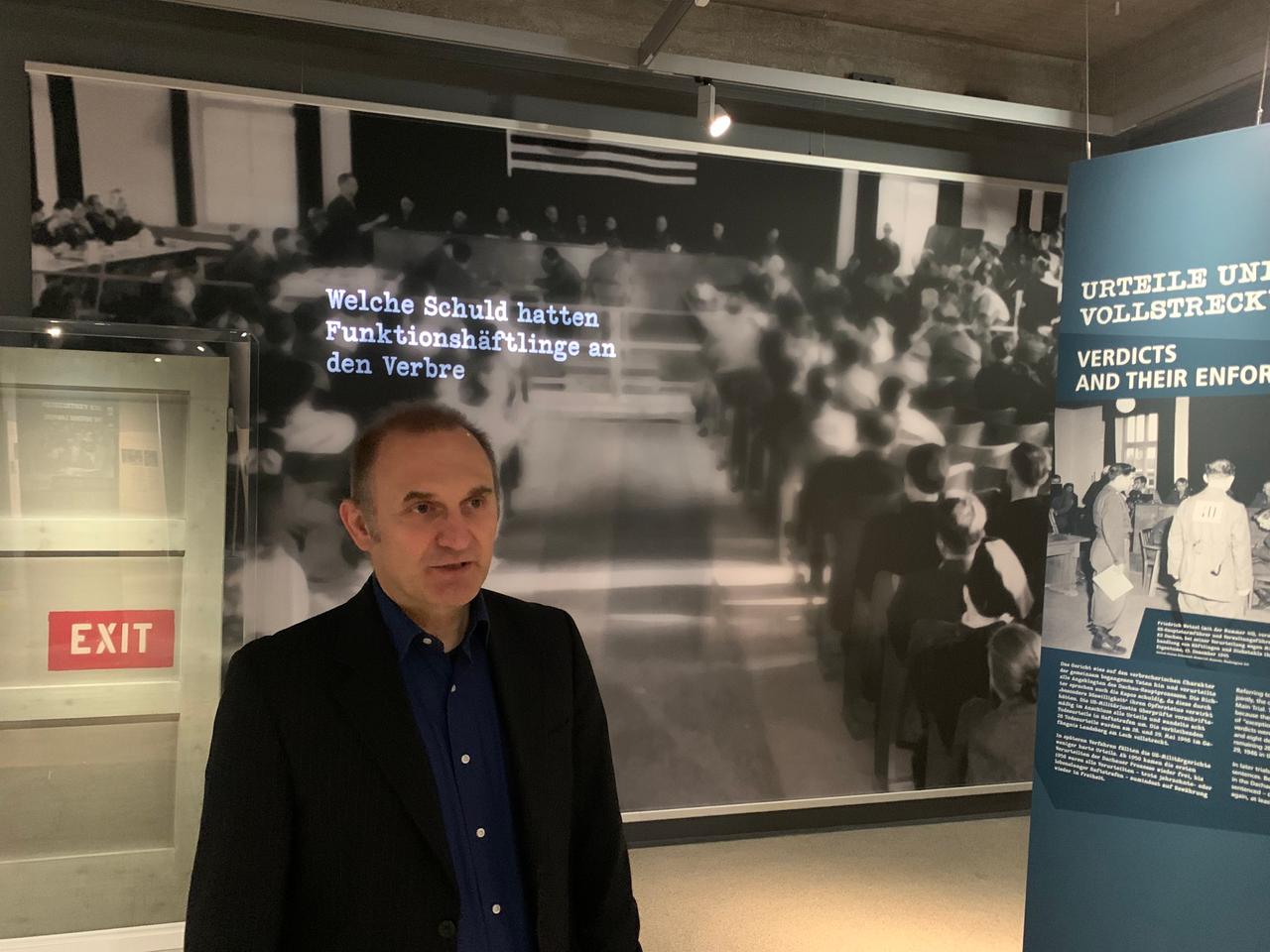 Ein Mann in dunklem Hemd und Jackett steht vor einer großen Fototafel mit einem unvollständigen Schriftzug. Es ist Christoph Thonwald, Leiter der Wissenschaftlichen Abteilung der Gedenkstätte Dachau in Bayern. Ein Mann in dunklem Hemd und Jackett steht vor einer großen Fototafel mit einem unvollständigen Schriftzug. Es ist Christoph Thonwald, Leiter der Wissenschaftlichen Abteilung der Gedenkstätte Dachau in Bayern.