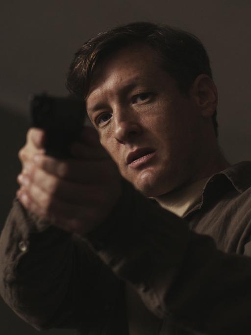 Ein Mann steht mit einer gezückten Pistole in einem dunklen Raum