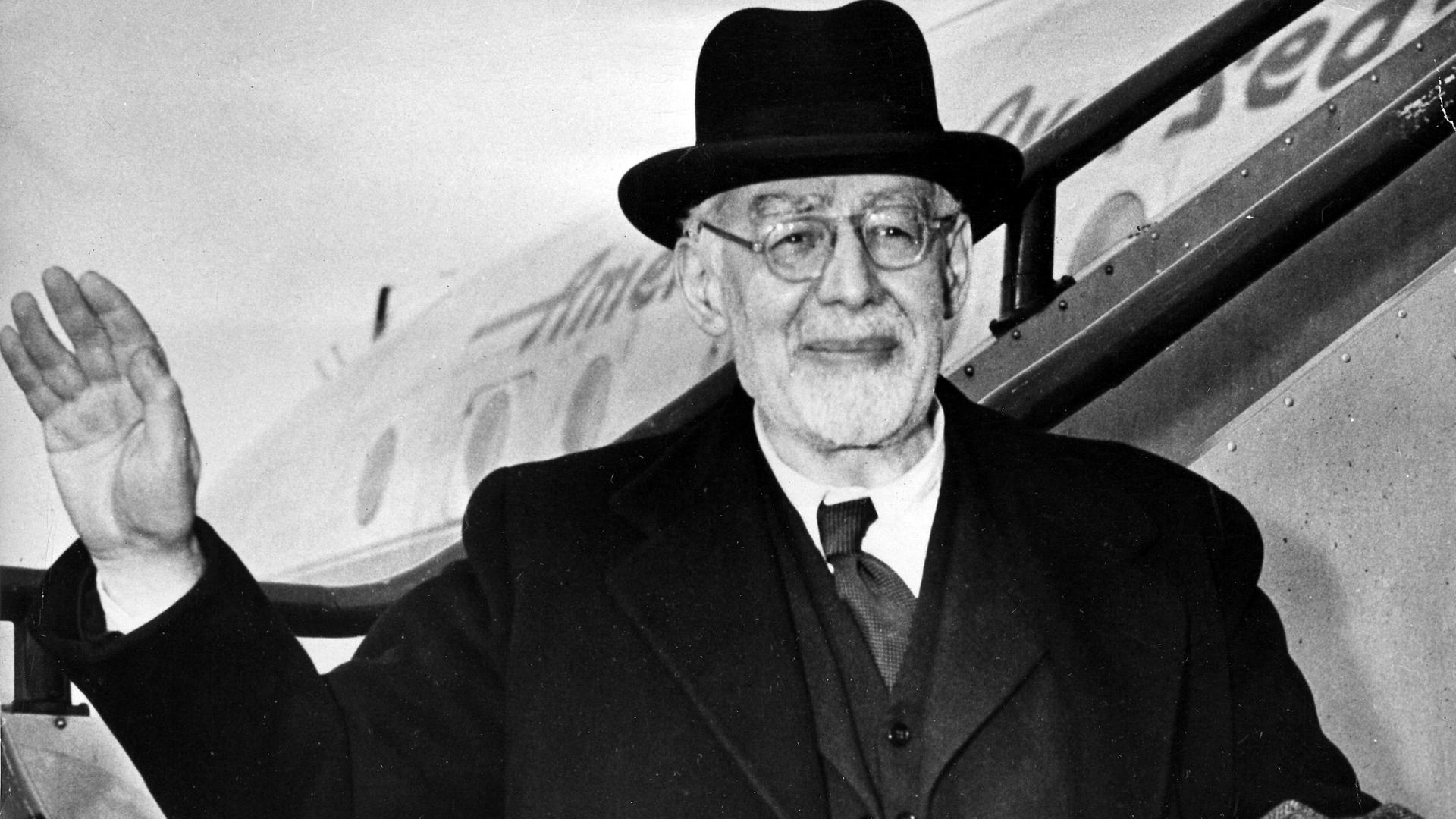 Eine Schwarzweiß-Fotografie zeigt einen älteren Mann mit schwarzem Hut, grüßend die Rolltreppe eines Flugzeugs herabsteigend Eine Schwarzweiß-Fotografie zeigt einen älteren Mann mit schwarzem Hut, grüßend die Rolltreppe eines Flugzeugs herabsteigend