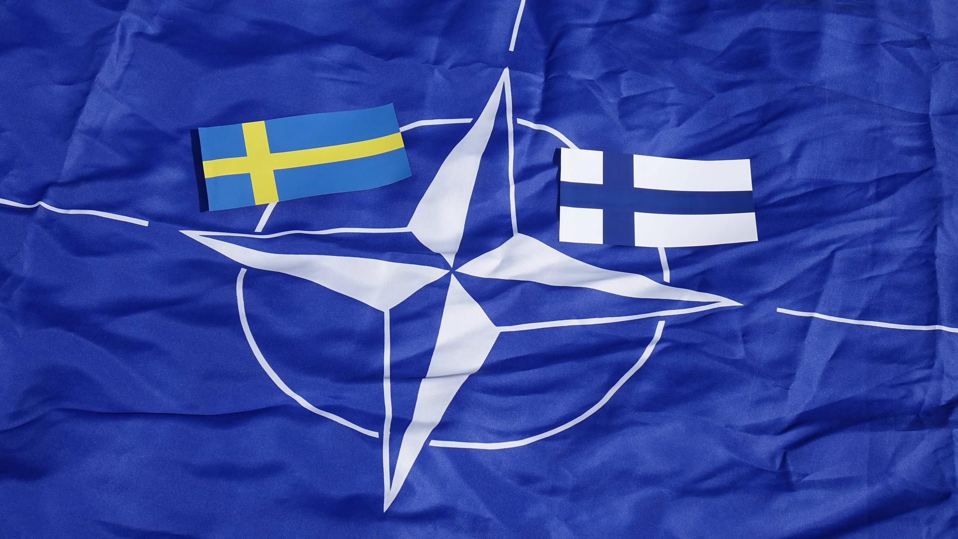 Kleine Flaggen von Schweden nund Finnland laigen auf einer großen Flagge der mit dem NATO-Logo. Kleine Flaggen von Schweden nund Finnland laigen auf einer großen Flagge der mit dem NATO-Logo.