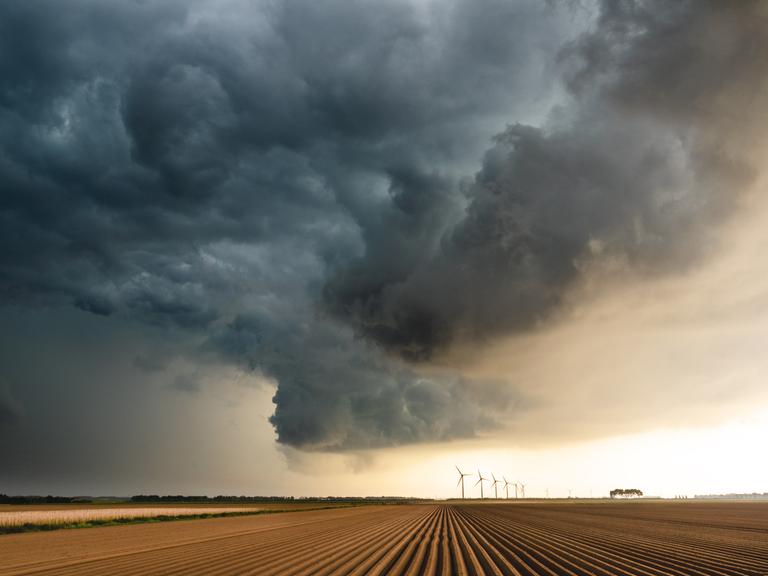Gewitterwolken über einem Feld. Im Hintergrund sind Windräder zu sehen, während die Sonne langsam durch die Wolken bricht.