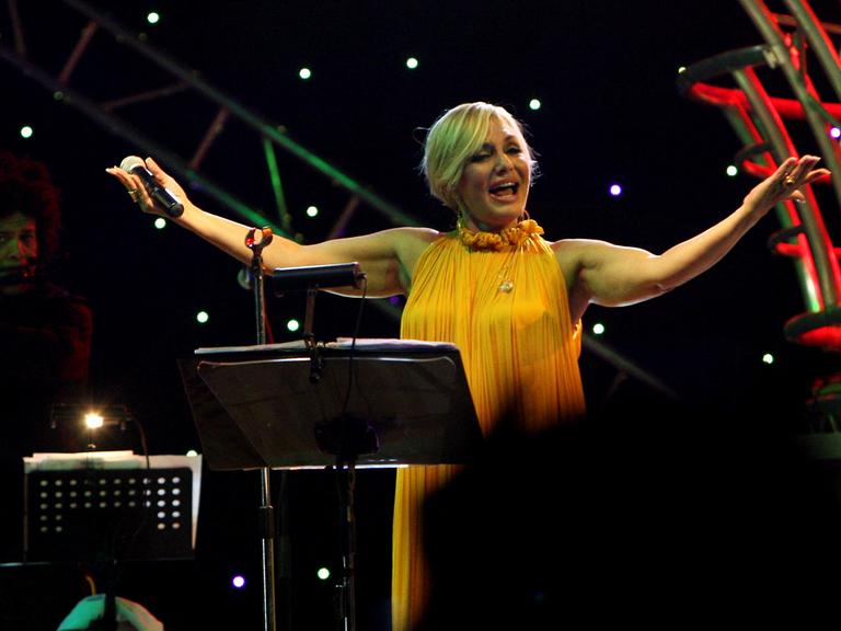 Die iranische Sängerin Googoosh steht singend auf einer Bühne und breitet die Arme aus.